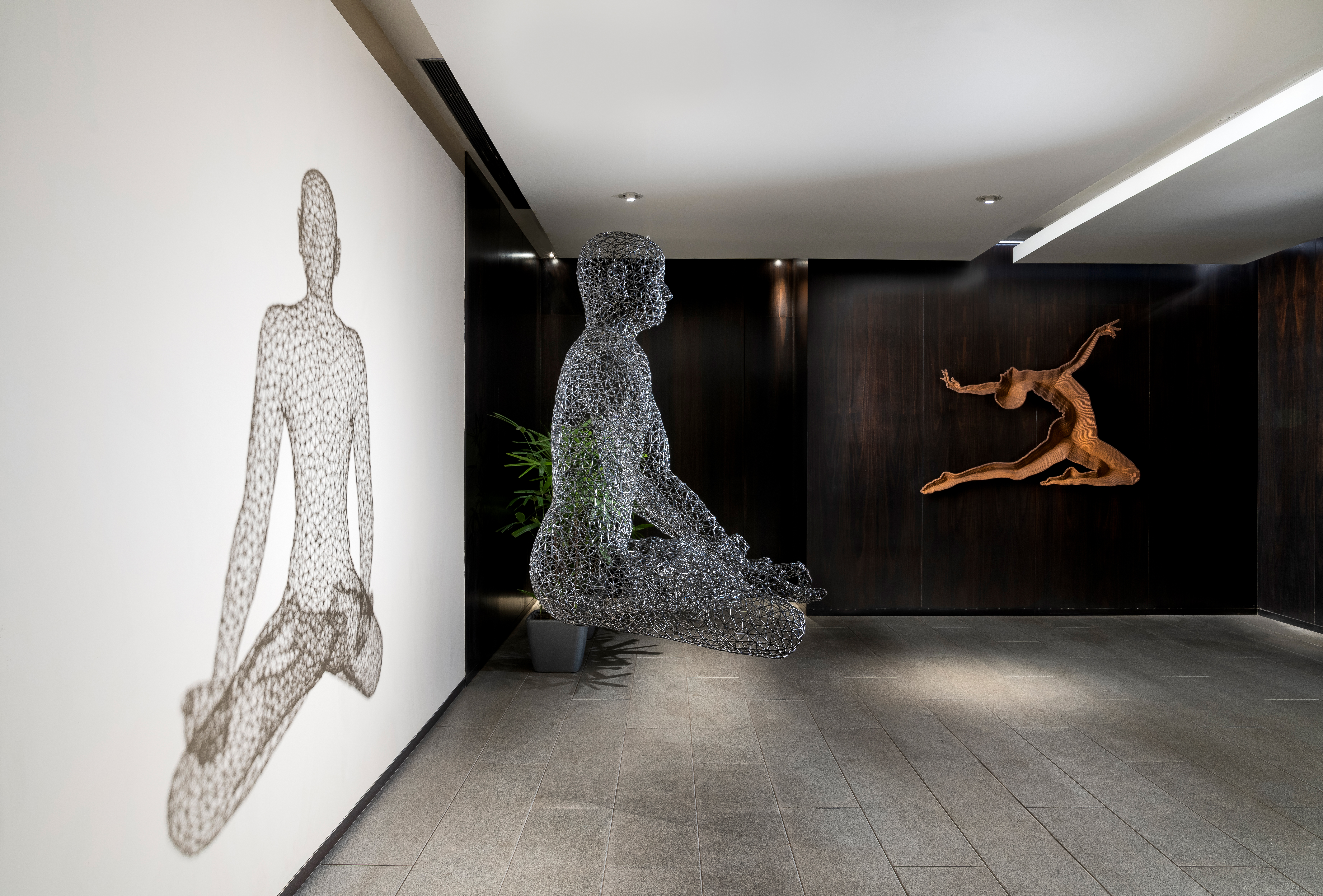 A gallery corridor with a floating wire-mesh meditation figure and a carved wooden dancer — Artemist Consultancy